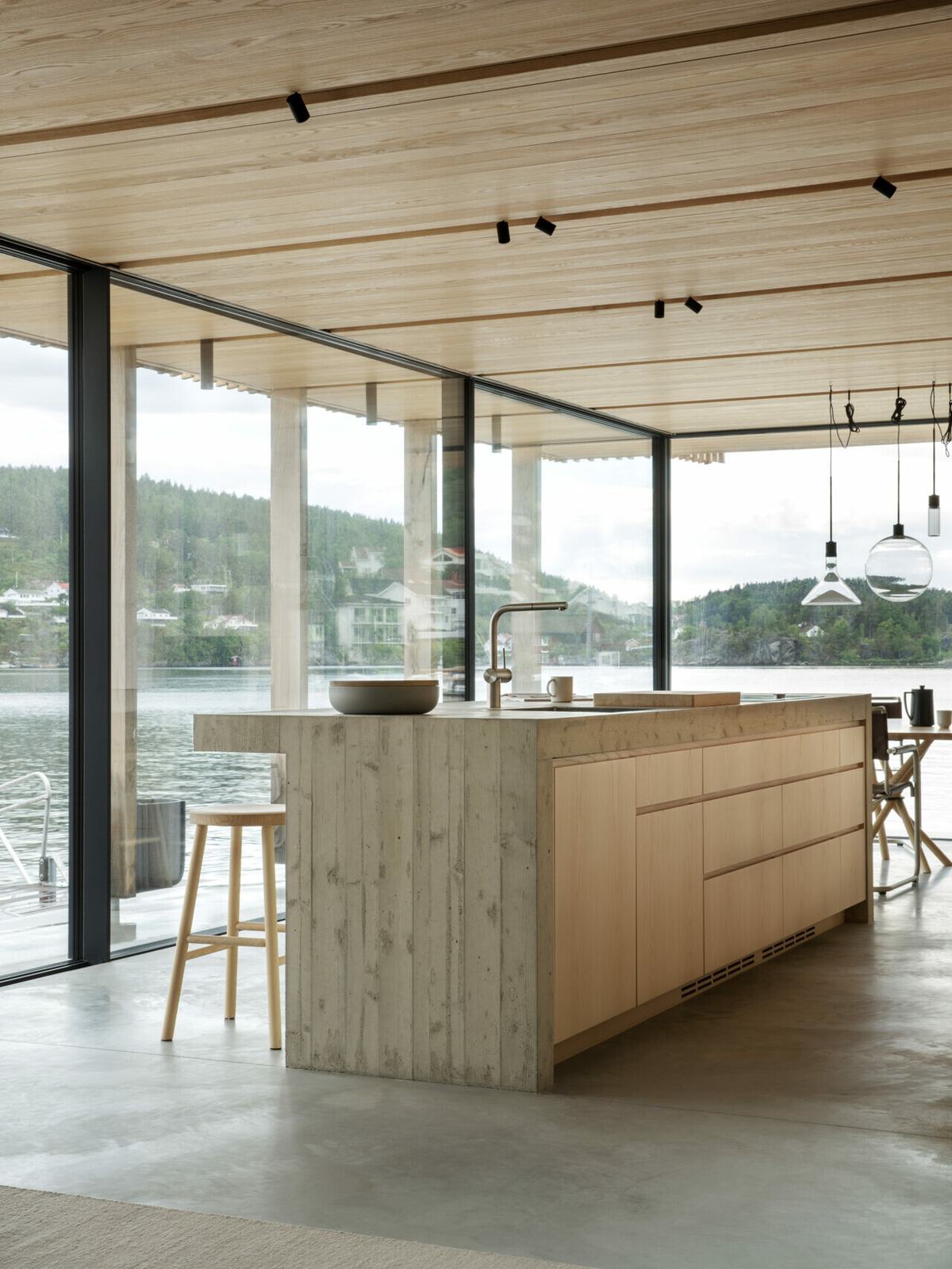 Kjøkkenøya i heltre strekker seg mot store glassfelt med utsikt over vannet. Skuffefrontene har freste grep, og benkeplaten i betong møter treverket i en rett linje.